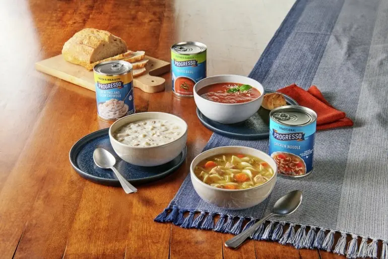 Bowls of Progresso soup, bread, and soup cans on wood table with blue table cloth.