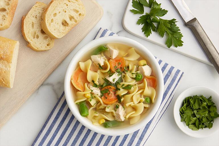 A bowl of chicken noodle Soup on a table next to bread and some herbs