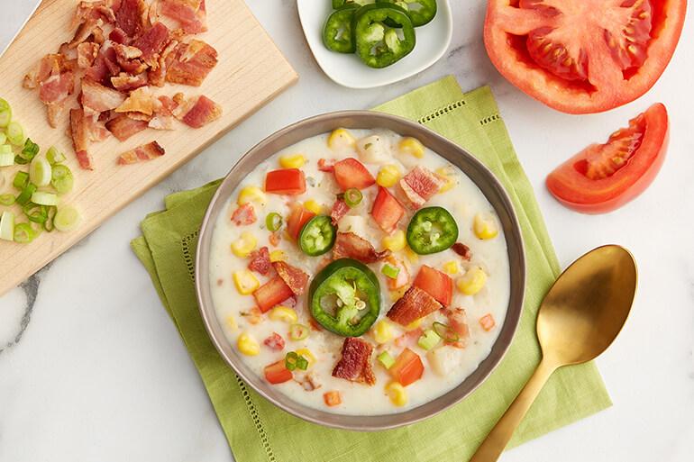A bowl of creamy corn chowder soup on a table next to a spoon and ingredients