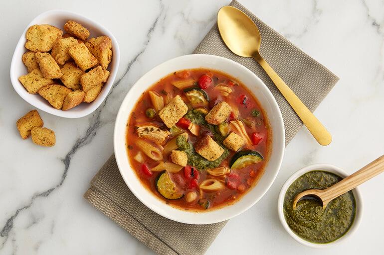A bowl of minestrone soup on a table next to ingredients and a spoon