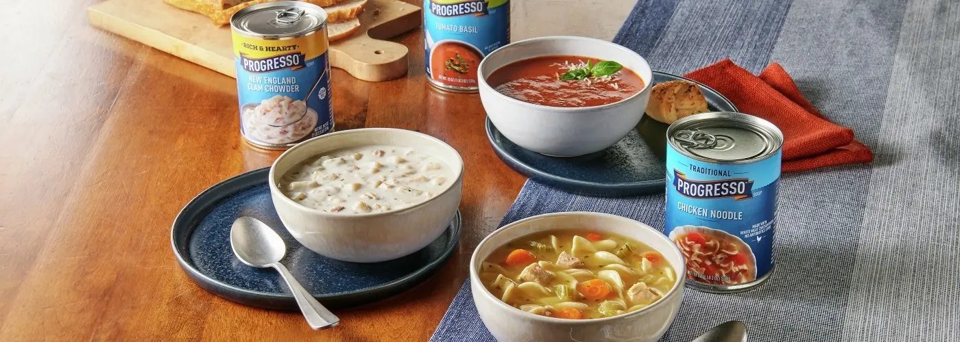 Bowls of Progresso soup, bread, and soup cans on wood table with blue table cloth.