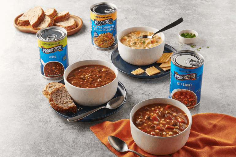 Three bowls of soup placed on a table, each accompanied by an open can of Progresso soup. The soups are Lentil, Chicken Pot Pie Style with Dumplings, and Beef Barley. Slices of bread, crackers, and a small bowl of chopped herbs are also on the table.